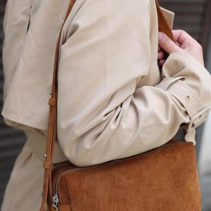 Pu&ograve; includere: Una borsa a tracolla rettangolare in camoscio marrone con chiusura a zip argentata e tracolla regolabile abbinata, indossata sopra un trench beige. La borsa presenta un design strutturato con cuciture visibili e dettagli in metallo.