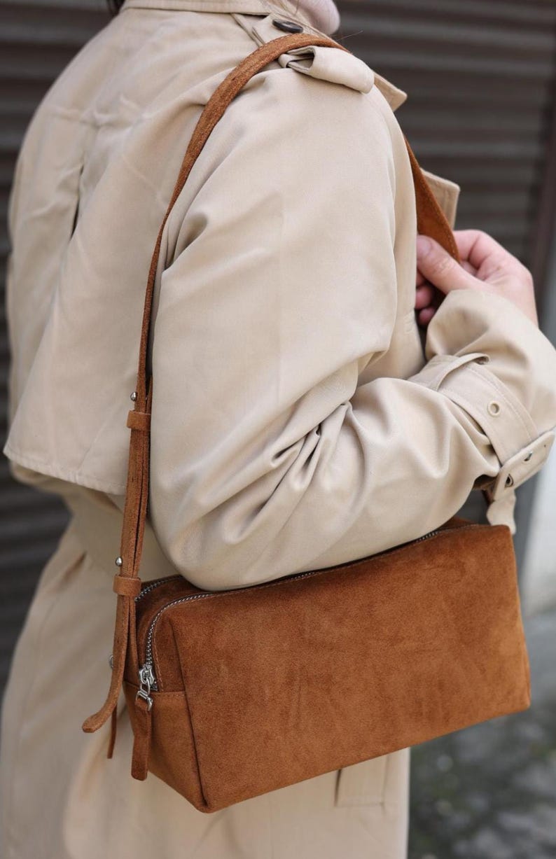 Pu&ograve; includere: Una borsa a tracolla rettangolare in camoscio marrone con chiusura a zip argentata e tracolla regolabile abbinata, indossata sopra un trench beige. La borsa presenta un design strutturato con cuciture visibili e dettagli in metallo.