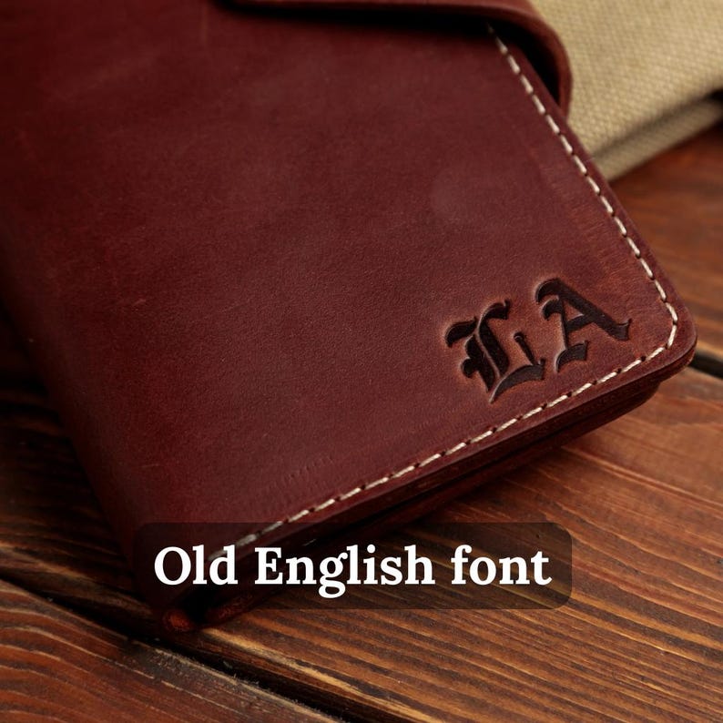 Puede incluir: Una cartera de cuero color burdeos oscuro con costuras blancas en contraste a lo largo de los bordes. La superficie est&aacute; grabada con las letras LA en una fuente Old English. El cuero tiene un acabado mate y una textura visible.