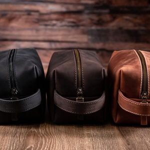 May include: Three rectangular toiletry bags in black, dark brown, and light brown leather. Each bag features a top zip, a side handle embossed with the text Lion Leather co., and visible stitching along the edges and handle attachments.