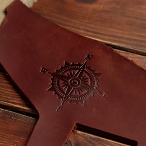 Puede incluir: Una pieza de cuero marr&oacute;n oscuro con un dise&ntilde;o de rosa de los vientos grabado en el centro. La br&uacute;jula incluye las letras N, E, S y W de los puntos cardinales en las puntas de la estrella, con un borde circular y l&iacute;neas radiantes.
