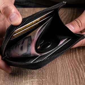 May include: A black zipped wallet featuring a main compartment containing folded paper currency, a small metal ring, and multiple credit cards stored in interior slots. The wallet is constructed with a dark fabric exterior and a matching interior lining.