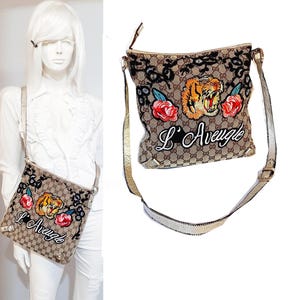 May include: A brown and beige canvas shoulder bag with a gold chain strap. The bag has an embroidered tiger face, flowers, and the text "L'Aveugle".