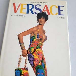 May include: A hardback book titled Versace by Richard Martin, featuring a cover image of a model wearing a colourful, sleeveless dress with a repeating pop-art face print. The model also wears a matching wide bracelet and carries a small handbag.