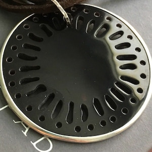 Könnte beinhalten: Ein schwarzer Anhänger mit silbernem Rand und einem ausgeschnittenen Design. Der Anhänger ist an einem braunen Lederband befestigt.