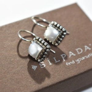May include: A pair of silver earrings with square, white pearl centers and beaded edges, resting on a brown box. The box has the brand name "SILPADA" and the tagline "find yourself" printed on it.