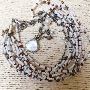 May include: Multi-strand beaded bracelet with white, brown, and gray beads. The bracelet features a silver charm with a white stone and a silver clasp.