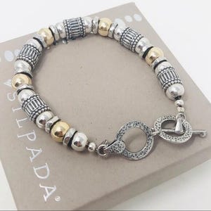 Op de afbeelding: Zilveren en gouden kralenarmband met een tuimelsluiting. De armband heeft een mix van zilveren en goudkleurige kralen, waaronder ronde en cilindrische vormen. De armband ligt op een lichtbruine doos met het woord "SILPADA" erop.