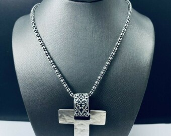 Könnte beinhalten: Eine silberne Kettenkette mit einem großen silbernen Kreuzanhänger. Das Kreuz hat eine strukturierte Oberfläche und ein detailliertes Design an der Oberseite.