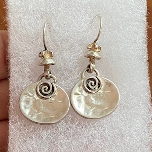 May include: A pair of silver earrings with a spiral design. Each earring features a circular pendant with a hammered texture and a spiral detail. The earrings have a hook closure and a decorative top. The earrings are displayed on a white surface.