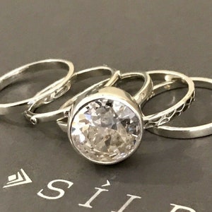 May include: A set of five silver rings with a large clear crystal stone in the center. The rings are stacked on top of each other.