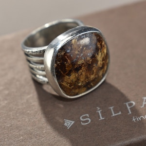 May include: A silver ring with a large, square-shaped stone. The stone has a mix of brown and gold tones. The ring's band has a textured design. The ring is on a brown surface.