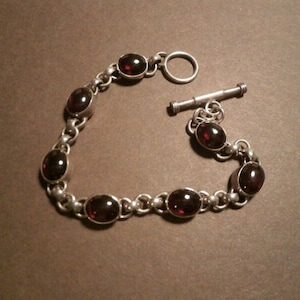 May include: Silver bracelet with oval, dark red gemstones. The bracelet has a toggle clasp and is made of interconnected links. The gemstones are set in individual bezels, creating a classic design.