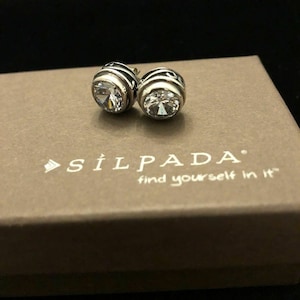 Pode incluir: Um par de brincos de prata com pedras de cristal transparentes engastadas em um design preto e prateado. Os brincos estão em uma caixa com o texto "Silpada find yourself in it".