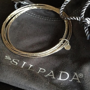 Puede incluir: Un brazalete de plata con múltiples bandas finas, descansando sobre una bolsa de terciopelo gris con la palabra "SILPADA" impresa en ella.