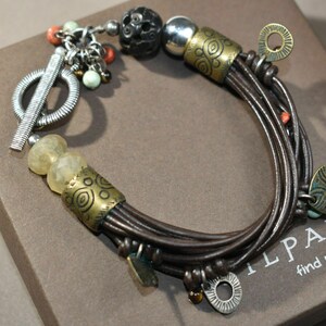 May include: A brown leather bracelet with multiple strands and various charms, including a round silver charm, a small yellow stone, and a silver toggle clasp. The bracelet also features a small, round, black bead and a larger, round, brown bead with a carved design. The bracelet is on a brown box with the text "ILPADA. find yourself in it" printed on it.