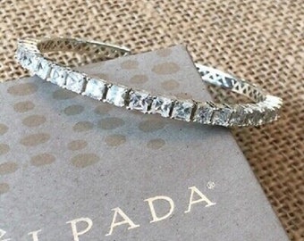 May include: A silver bangle bracelet with a row of clear square-shaped stones. The bracelet is on a gray box with the word "ILPADA" printed on it.
