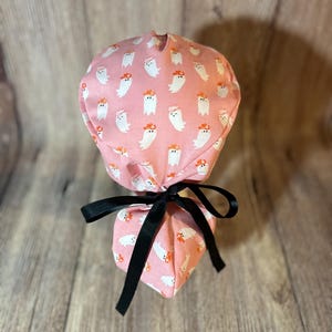 May include: Pink surgical scrub hat with a pattern of white ghosts wearing orange hats. The hat is tied with a black ribbon.