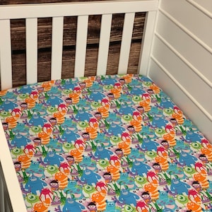 May include: A white crib with a fitted sheet featuring a colorful pattern of various characters from the Disney Pixar movie Monsters Inc. The sheet is covered in a repeating pattern of blue, green, orange, and purple monsters.