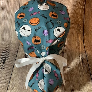 May include: A teal scrub cap featuring a Halloween-themed pattern. The design includes Jack Skellington faces, orange pumpkins, bats, and candy. The cap is tied with a white ribbon.