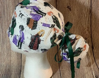 May include: A white scrub cap featuring a repeating pattern of illustrated characters, tombstones, and black splatters. The cap includes a dark green ribbon tie at the back to secure the fabric, which is gathered to accommodate hair of various lengths.