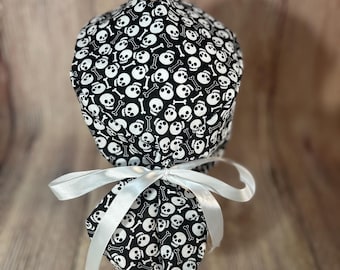 May include: A black and white skull print scrub cap with a white ribbon. The cap is made of fabric and features a repeating pattern of white skulls and crossbones on a black background. The cap is tied with a white satin ribbon.