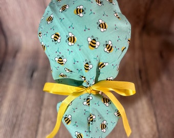 May include: A light teal scrub cap with a repeating pattern of yellow and black bees and small purple berry sprigs. The cap is tied with a bright yellow ribbon bow. This cap is designed for medical professionals.