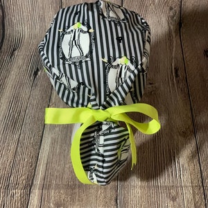 May include: A black and white striped scrub cap with a neon yellow bow. The cap features a repeating design of a character from the movie Beetlejuice. The cap is made of fabric and is tied with a bow.
