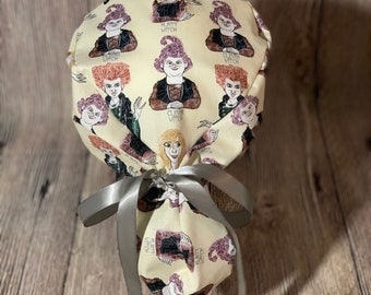 May include: A cream-colored scrub cap featuring a repeating pattern of cartoon witches in various poses. The cap is tied with a gray satin ribbon. The witches have different hair colors and are wearing dark clothing.