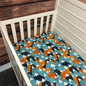 May include: A white crib with a fitted sheet featuring a geometric mountain pattern in shades of blue, orange, and white.