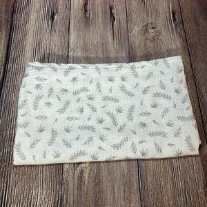 May include: A white muslin swaddle blanket with a delicate green leaf pattern. The blanket is folded in half and lying on a wooden surface.