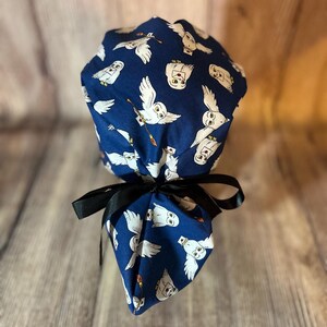 May include: A dark blue scrub cap featuring a repeating pattern of white owls carrying letters and brooms. The cap is tied with a black ribbon. The owls are depicted in flight, with detailed facial features and holding letters.