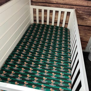 May include: A white crib with a green and black fitted sheet featuring a repeating pattern of the Milwaukee Bucks logo.