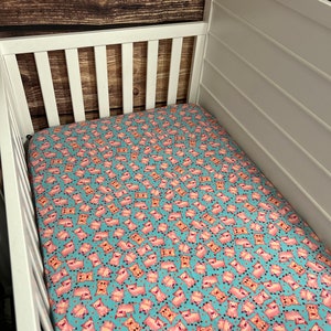May include: A turquoise and pink crib sheet with a pattern of cartoon pigs. The pigs are pink with black eyes and snouts. The sheet is made of soft, breathable fabric.