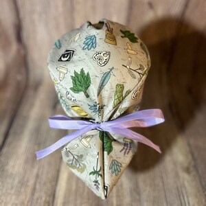 May include: A light gray scrub cap with a repeating pattern of crystals, leaves, and other mystical symbols. The cap is tied with a lavender ribbon. The fabric has a vintage, slightly distressed look.