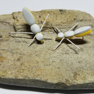 May include: Two white foam fly fishing lures with yellow accents on a light brown rock. The lures have white legs and are designed to resemble insects.