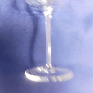 May include: A clear, stemmed glass with a round base and a long, slender stem. The glass is made of a transparent material, reflecting light. The background is a solid blue colour.