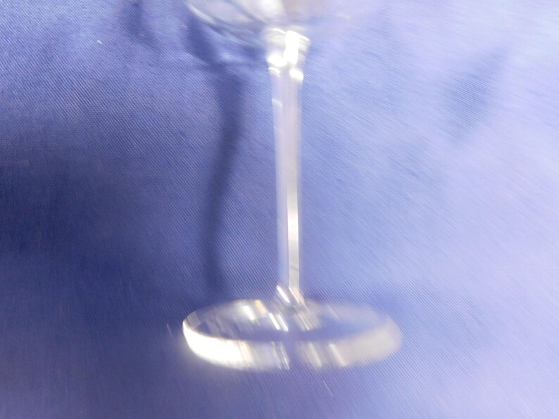 May include: A clear, stemmed glass with a round base and a long, slender stem. The glass is made of a transparent material, reflecting light. The background is a solid blue colour.
