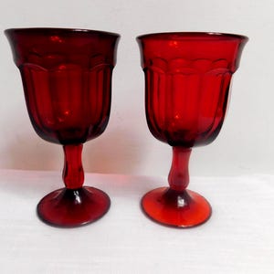 May include: Two matching deep red glass goblets featuring a vertical ribbed pattern along the bowl and a scalloped rim design. Each glass stands on a sturdy pedestal base with a smooth stem, showcasing a translucent ruby color throughout the glass.
