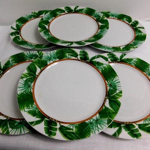 May include: Six white ceramic plates with a green and brown tropical leaf design around the rim. The plates are arranged in a circular pattern, showcasing the decorative border.