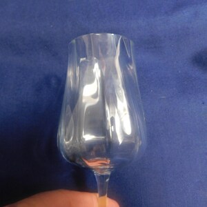 May include: A clear wine glass with a tulip-shaped bowl and vertical ridges. The glass is held against a blue background. The stem is thin and the glass is empty.