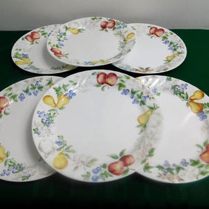 May include: Six white ceramic plates with a decorative border of red apples, yellow pears, and blue flowers. The plates are arranged on a dark green surface, showcasing a classic fruit design.