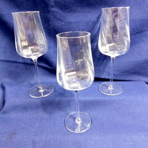 May include: Three clear wine glasses with a distinctive, curved shape. Each glass features a long, thin stem and a broad, angled bowl. The glasses are made of transparent glass, allowing the contents to be seen. The glasses are set against a blue background.