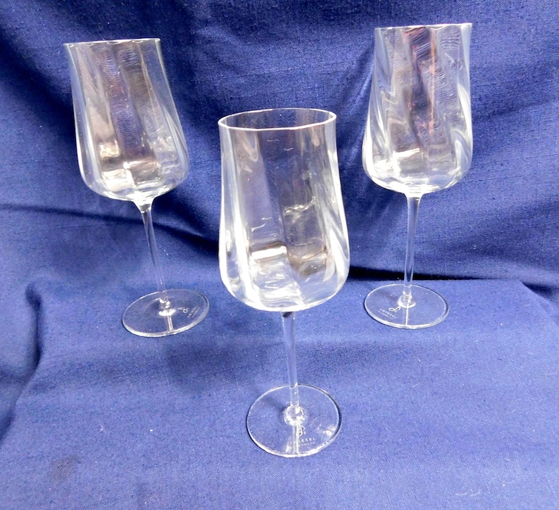 May include: Three clear wine glasses with a distinctive, curved shape. Each glass features a long, thin stem and a broad, angled bowl. The glasses are made of transparent glass, allowing the contents to be seen. The glasses are set against a blue background.