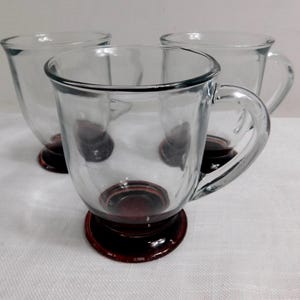 May include: Three clear glass mugs with a dark red base and handle. The mugs are empty and sit on a white surface. The glass has a slight swirl design. The mugs are suitable for serving hot beverages.