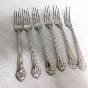 May include: Six silver forks with a decorative, scalloped design on the handles. The forks are arranged on a white, textured surface. The tines of the forks are visible.