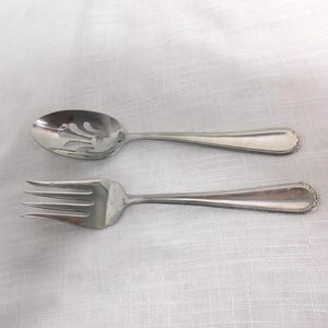 May include: A stainless steel slotted spoon and a fork are displayed on a white surface. Both utensils have a decorative rope-like design on the handles and a classic, elegant appearance. The spoon has a slotted bowl.