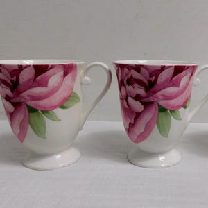 May include: Two white ceramic mugs with a floral design. The mugs feature a large pink peony flower with green leaves. The mugs have a curved handle and a pedestal base. The mugs are on a white surface.