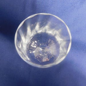 May include: A clear glass tumbler with a wavy rim, viewed from above. The glass has a faceted interior and sits on a blue surface. The transparent design allows light to pass through, showcasing the glassware's details.