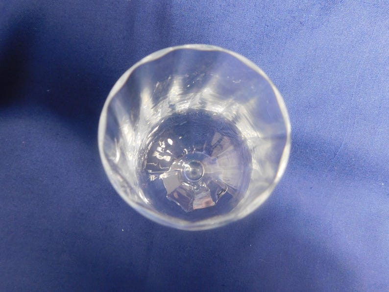 May include: A clear glass tumbler with a wavy rim, viewed from above. The glass has a faceted interior and sits on a blue surface. The transparent design allows light to pass through, showcasing the glassware's details.